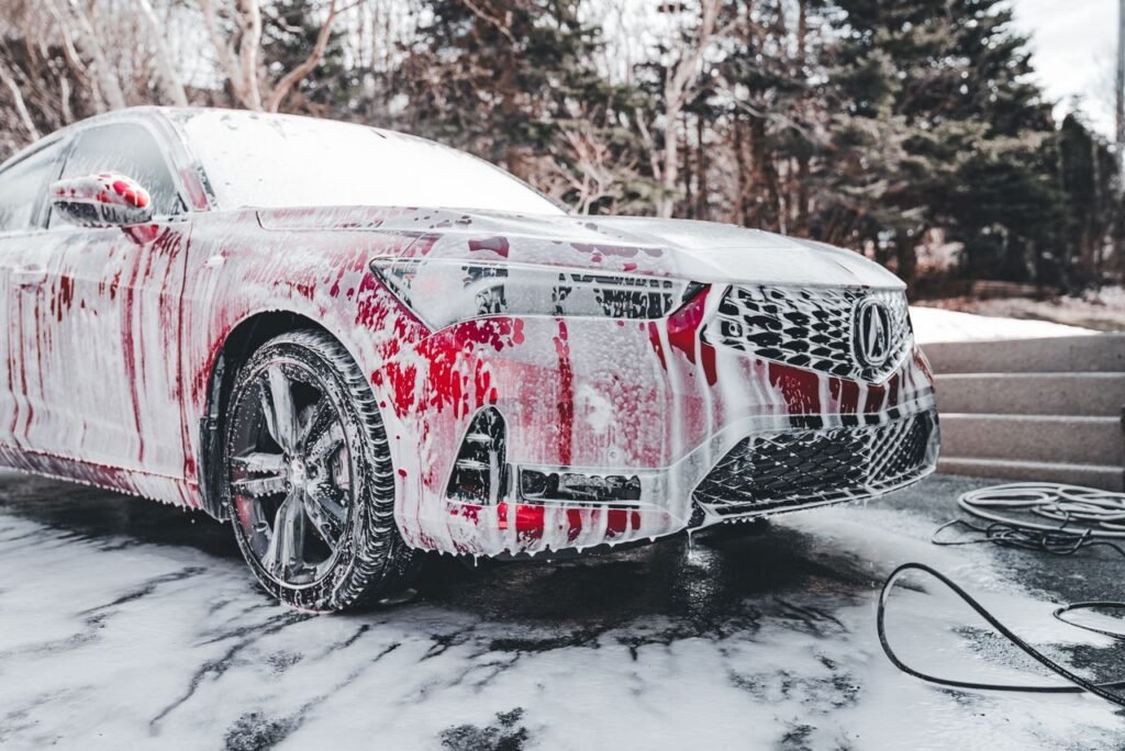 A vibrant red car covered in soapy foam during a thorough outdoor wash under daylight.